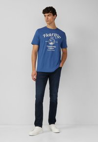 Niebieska bawełniana koszulka z białym nadrukiem tekstu, okrągły dekolt, krótkie rękawy, w połączeniu z ciemnymi dżinsami i białymi trampkami.