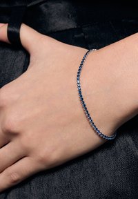 Srebrna bransoletka z niebieskimi kamieniami szlachetnymi, o delikatnym wzornictwie, które obejmuje nadgarstek. Gładka tekstura, na ciemnym tle.