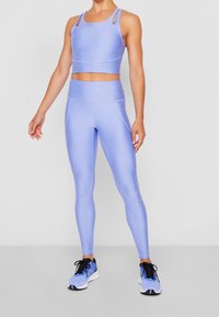 Žena v světlých fialových sportovních legínách, odpovídající zkrácené halence a modrých běžeckých botách, stojící na jednobarevném pozadí.
