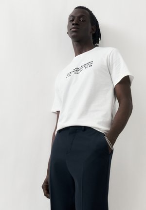 Άνδρας με dreadlocks που φοράει λευκό T-shirt Calvin Klein και σκούρο παντελόνι, στέκεται με το ένα χέρι στην τσέπη μπροστά σε απλό φόντο.