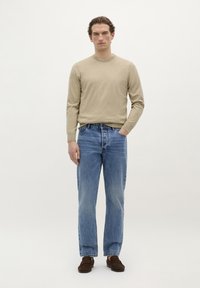 Pulover tricotat bej, blugi din denim ușor bleu, loafers maro. Modelul stă drept, cu mâinile în buzunare, pe un fundal alb simplu.