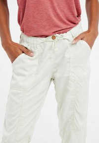 Pantaloni cargo di colore chiaro con una consistenza morbida, vita elasticizzata, chiusura con cordino e tasche laterali. Presentano un dettaglio con bottone sulla vita.