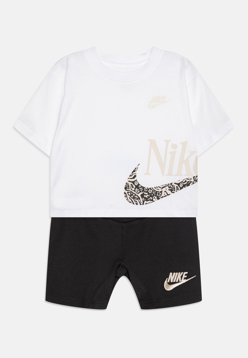 Λευκή κοντομάνικη μπλούζα με μπεζ λογότυπα της Nike και μαύρες διακοσμητικές λεπτομέρειες, συνδυασμένη με μαύρες ριγέ σορτς που έχουν μπεζ λογότυπο Nike.
