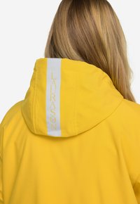 Keltainen hupparitakki, jossa on valkoinen pystysuora raita ja selässä teksti "LAURASØN" elegantilla fontilla. Silkkinen, kevyt kangas.