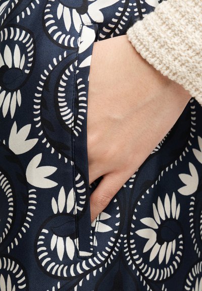 Χέρι με μπεζ πλεκτό μανίκι που εισέρχεται στην πλαϊνή τσέπη σκουρόχρωμου μπλε υφάσματος με λευκό και μαύρο φλοράλ μοτίβο πασλέι.