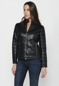 Veste en cuir noir au design ajusté. Comprend des panneaux matelassés, un col haut et des poches zippées. Portée sur un jean en denim bleu.