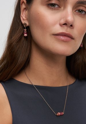 Frau mit hängenden Ohrringen und einer Halskette aus pinken und rötlichen Perlen, gekleidet in einem dunklen, ärmellosen Oberteil.