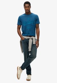 Camiseta de algodón azul con un estampado gráfico, suéter gris claro atado a la cintura, jeans oscuros y zapatillas beige con acentos marrones.