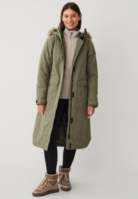 Larga chaqueta parka verde oliva con capucha forrada de piel, cierres ajustables, bolsillos y un corte holgado, llevada sobre un suéter beige y leggings negros.