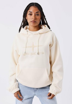Donna con capelli intrecciati che indossa un hoodie color crema con un logo a stella e jeans azzurri chiari, in piedi contro uno sfondo bianco uniforme.