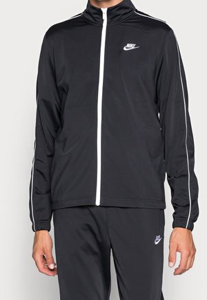 Mand iført sort Nike træningsdragt med hvide striber, lynlåstrøje foran og matchende bukser med Nike-logoer på bryst og lår.