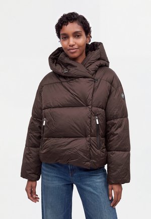 Modèle portant une doudoune matelassée marron foncé à capuche oversize et un jean bleu, sur un fond blanc uni.