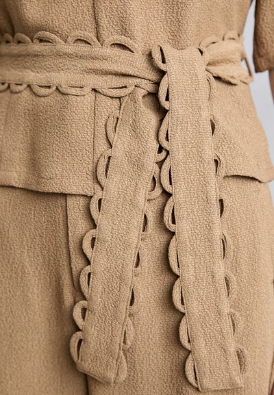 Beige textured fabric with a scalloped trim belt tied at the waist. The design features a layered appearance and soft texture.