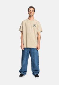 Beige bavlněné tričko s krátkým rukávem a kruhovým logem, spárované s volnými modrými džíny a černými botami na slip-on.