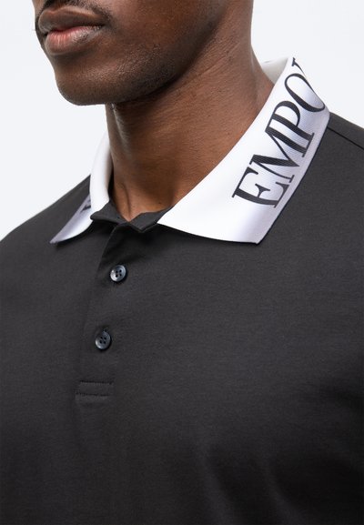 Κοντινό πλάνο ενός άνδρα που φοράει μαύρο πόλο μπλουζάκι με δύο κουμπιά και λευκό γιακά με μεγάλο μαύρο κείμενο «EMPOR».