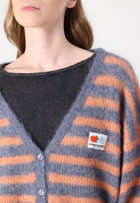 Randig kofta i en blandning av blå och orange mohair, med V-hals, knappstängning och en logotyp patch på vänster bröst. Mjuk, texturerad tyg.