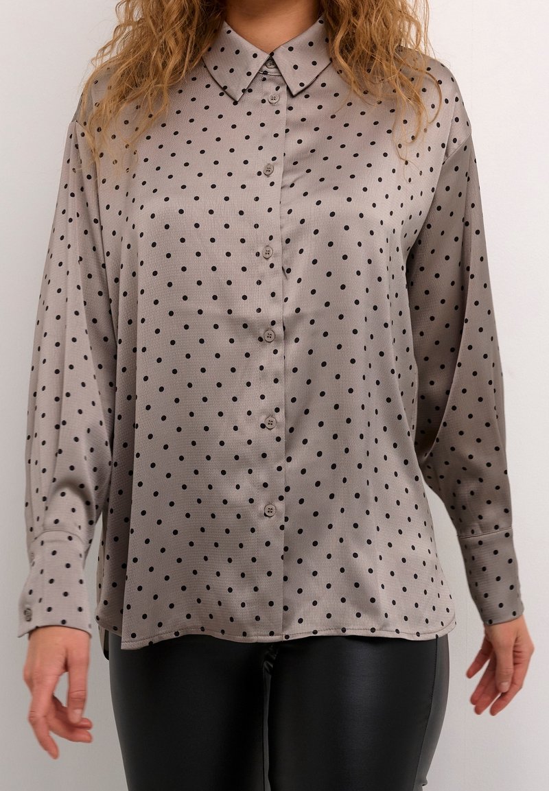 Femme portant une chemise en satin grise à pois noirs boutonnée et un pantalon en cuir noir, debout devant un fond uni.