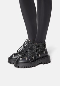 Botas negras de tobillo hechas de cuero texturizado con una suela de goma gruesa, que cuentan con cordones gruesos en blanco y negro y acentos de hardware metálico.