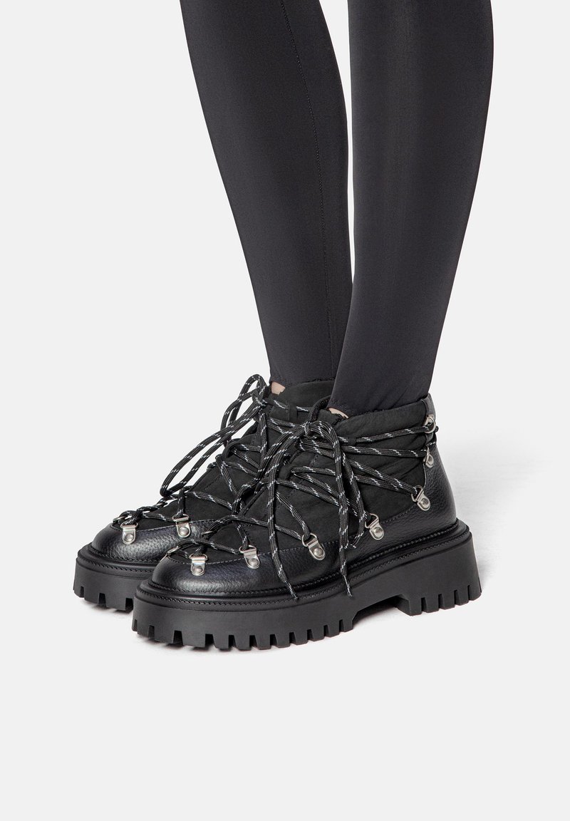 Botas negras de tobillo hechas de cuero texturizado con una suela de goma gruesa, que cuentan con cordones gruesos en blanco y negro y acentos de hardware metálico.