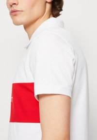Biała koszulka polo z czerwoną poziomą paskiem. Posiada klasyczny kołnierzyk, krótkie rękawy oraz małe haftowane logo na rękawie.