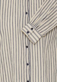 Krém színű, tengerészkék vízszintes csíkokkal díszített gombos ing, strukturált gallérral és hosszú ujjakkal, amelyek gombos mandzsettával záródnak.