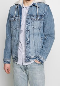 Veste en denim avec un délavage bleu clair, deux poches poitrine et une capuche doublée en fourrure. Présente des coutures contrastantes et un bouton de fermeture.