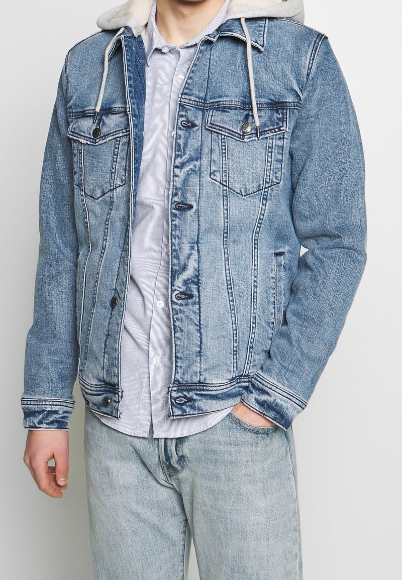 Veste en denim avec un délavage bleu clair, deux poches poitrine et une capuche doublée en fourrure. Présente des coutures contrastantes et un bouton de fermeture.
