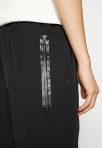 Pantalones negros con un bolsillo lateral con cremallera negra brillante y texturizada. La tela parece ligera y suave.