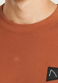 Tkanička v barvě rezavé, s kulatým výstřihem, krátkými rukávy a černou textilní visačkou s bílým designem na levé straně hrudi.