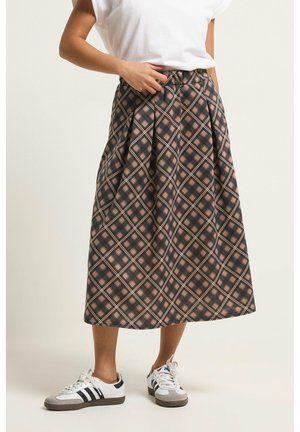 Kobieta w czarno-różowej w kratę midi spódnicy, białej koszulce i białych trampkach z czarnymi paskami, stojąca na jednolitym tle.