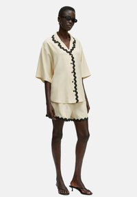 Beige shirts met korte mouwen en zwarte golvende afwerking en knopen, gecombineerd met bijpassende shorts met golvende randen, gemaakt van een gladde stof.