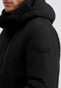 Schwarze isolierte Jacke mit hohem Kragen, verstellbare Kapuze und strukturierter Stoffoberfläche. Enthält ein kleines Logo-Patch am Ärmel.