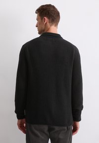 Black textured cardigan featuring a collared neckline and long sleeves. The fabric has a ribbed pattern and a relaxed fit.
