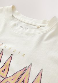 Cream-colored cotton t-shirt with a round neckline, featuring graphic designs in pink and black at the front. Text reads "MANIA."