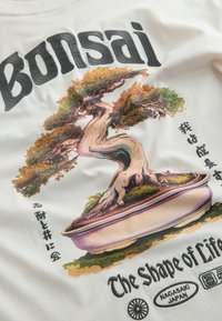 Wit T-shirt met een kleurrijke bonsai-boom graphic met een bruine stam en groene bladeren, geaccentueerd met Japanse tekst en "The Shape of Life."