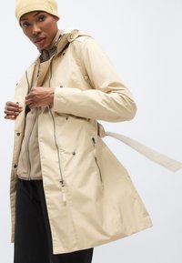 Μπεζ αδιάβροχο μπουφάν με κουκούλα, φερμουάρ μπροστά και κουμπώματα με πατιλέτα. Διαθέτει δύο πλαϊνές τσέπες και ζώνη με δέσιμο στη μέση.