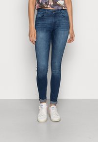 Jeans ajustados en denim azul oscuro, con una cintura de altura media, detalles en los bolsillos frontales y puños enrollados, combinados con zapatillas blancas.