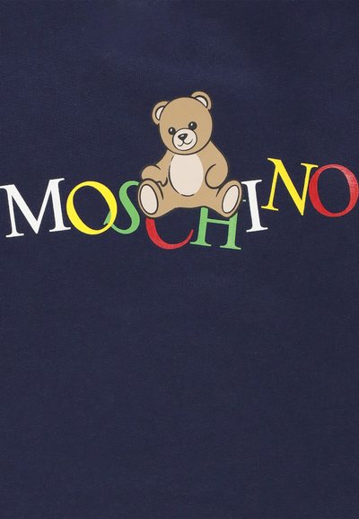 Un orsetto di peluche seduto sopra lettere colorate che formano la parola "MOSCHINO" su uno sfondo di tessuto scuro.