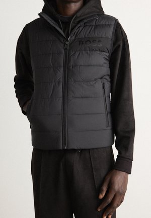 Man draagt een zwart doorgestikt vest met "BOSS" logo over een zwarte hoodie met lange mouwen en bijpassende broek, staand met één hand in de vestzak.