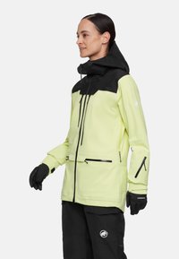 Helle gelbe wasserdichte Jacke mit schwarzen Akzenten, verstellbare Kapuze, Frontreißverschluss-Tasche und seitlichen Reißverschlusstaschen. Kombiniert mit schwarzen Hosen und Handschuhen.