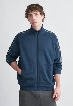 Marineblå zip-up jakke med høy krage, to frontlommer, elastiske mansjetter og en logo på brystet; har grå striper på ermene.