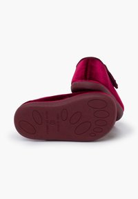 Zapatilla de terciopelo rosa con punta redondeada, acento de lazo en el mismo tono y suela de goma texturizada con patrones circulares. Fabricada en España.