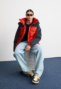 Rød og sort pufferjakke med hætte, parret med afslappede blå denimjeans og flerfarvede sneakers. Accessories inkluderer mørke solbriller.
