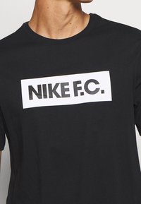 Sort bomulds T-shirt med en hvid rektangulær panel, der har teksten "NIKE F.C." i fed sort skrift. Korte ærmer og rund halsudskæring.