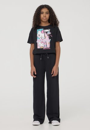 Maglietta nera a maniche corte con grafica e testo colorati di "Hello Kitty"; abbinata a pantaloni sportivi neri a gamba larga e scarpe da ginnastica bianche.