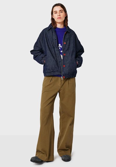 Person wearing a navy striped jacket with red buttons, a blue sweater, wide brown pants, and black shoes standing against a plain background.