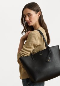 Μεγάλη μαύρη δερμάτινη τσάντα tote με υφή, δύο λουριά για τον ώμο και μια μικρή προσαρτημένη ετικέτα με χρυσά γράμματα.