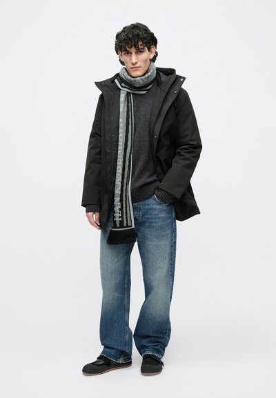 Parka impermeabilă neagră, pulover gri, blugi denim albaștri, eșarfă neagră cu model text, pantofi negri, stând în picioare pe un fundal simplu.