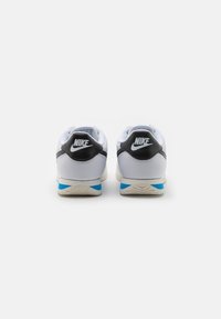 Fehér felsőrésszel, fekete sarokbetétekkel és világoskék részletekkel rendelkező Nike sportcipő, texturált mintával és logóval.