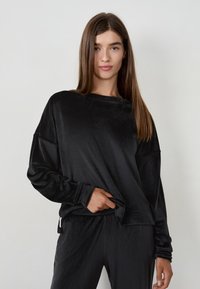 Giovane donna con lunghi capelli castani, vestita con una maglietta a maniche lunghe in velluto nero di taglio largo e pantaloni abbinati, in piedi contro uno sfondo chiaro e semplice.